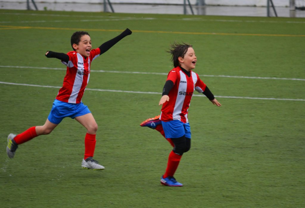 Two school aged boys celebrate by running down the soccer pitch together looking very excited in their matching uniforms.