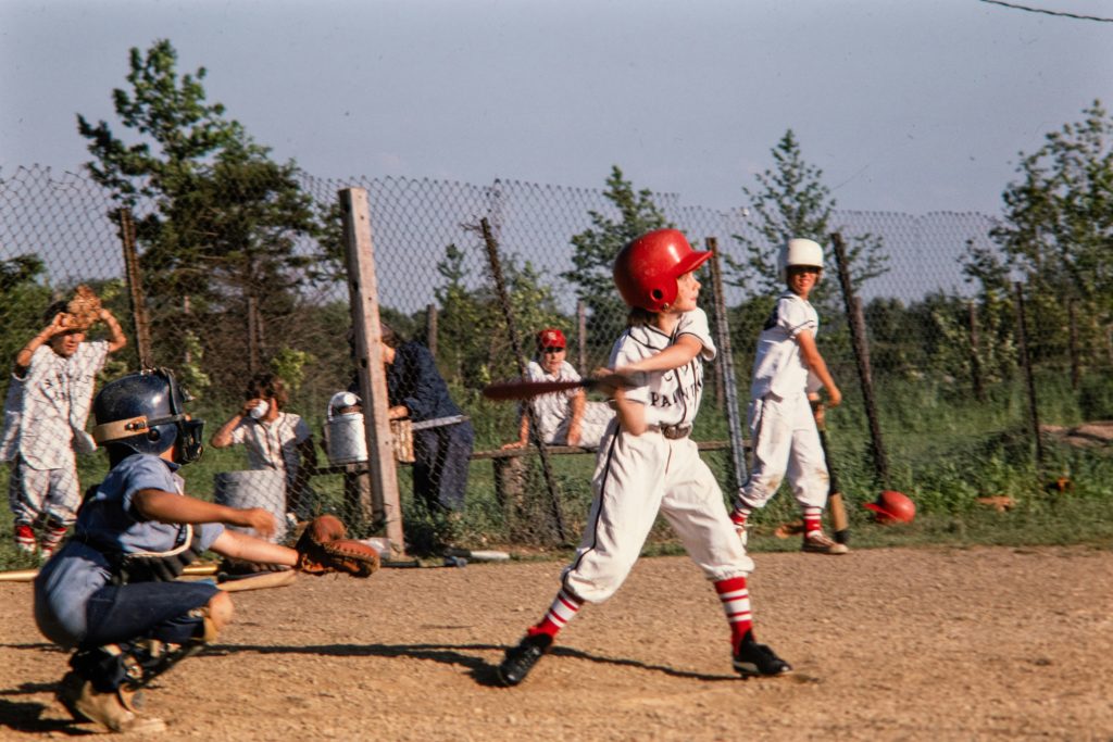 A catcher and a batter stand at home plate waiting for the pitch. The batter is mid swing. A group of children stand behind a fence watching them play baseball.