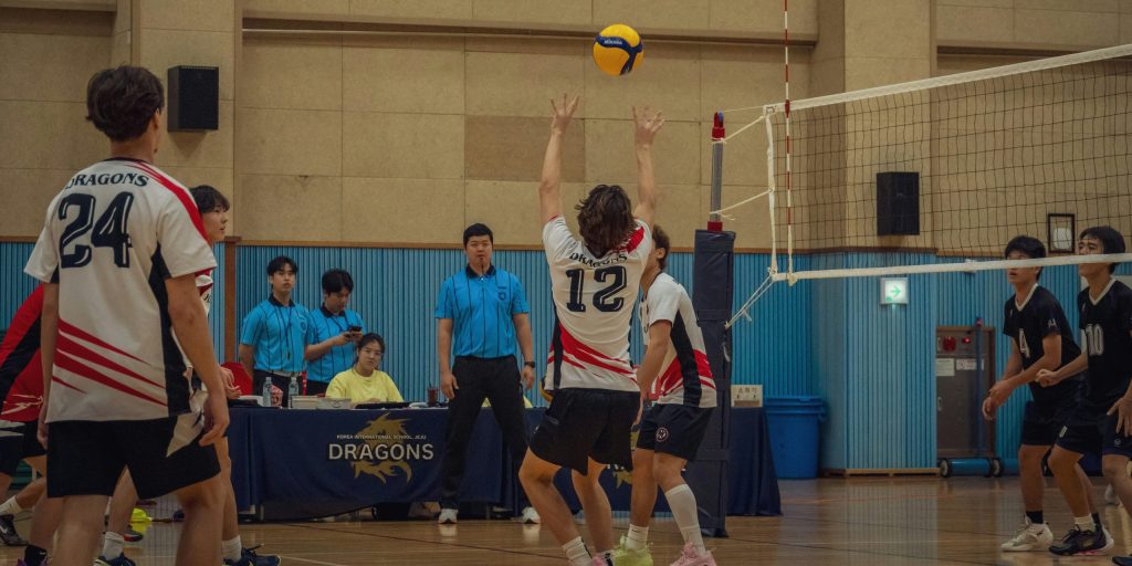Young teens play volley ball. The white team's setter prepares to receive the ball and set it for the spiker getting ready to run to the net. The white team's jerseys say 'Dragons'