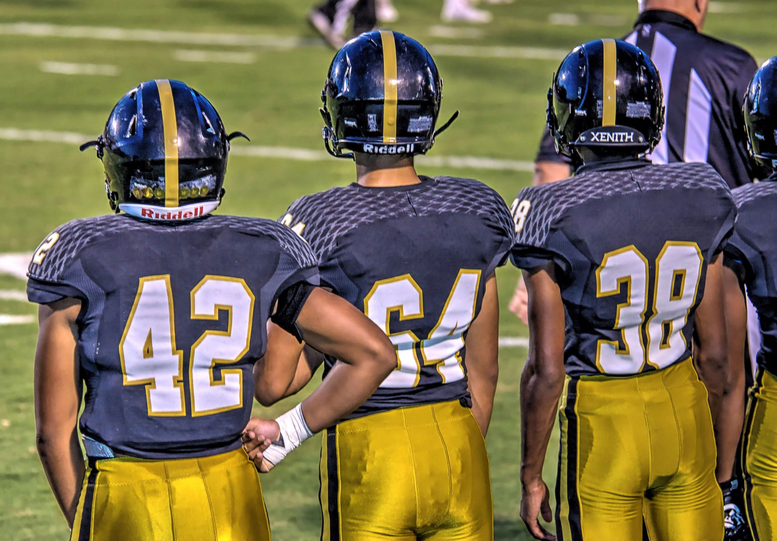 Teenaged American footballers stand in full uniform with helmets on and backs towards the camera on the sidelines of a field watching the current play.