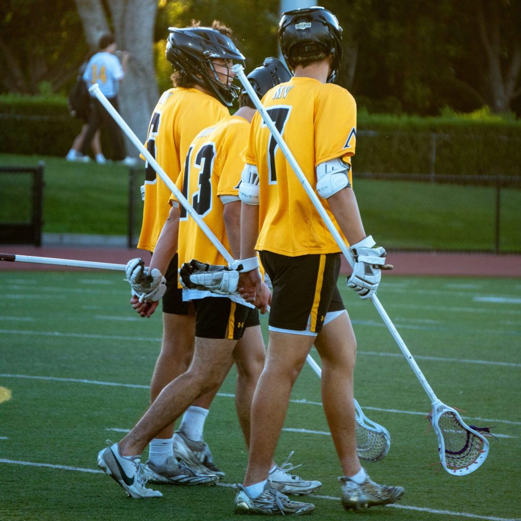 Three lacrosse players, two defenders and an attacker walk down the field together in matching uniforms.