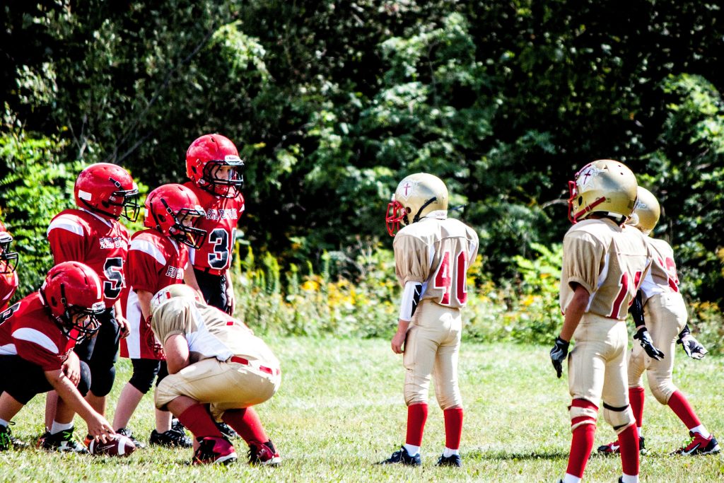 Young children playing American Football line up a the line of scrimmage to get ready for the next play. There is a red team and a gold team.